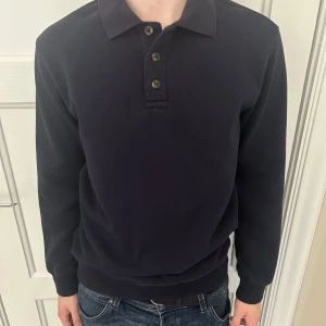 Mörkblå pikétröja från H&M - Säljer en stilren mörkblå pikétröja från H&M med klassisk krage och tre knappar framtill. Tröjan har långa ärmar och är tillverkad i ett mjukt material. 