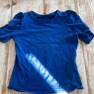 Blå topp med puffärmar från ONLY - Säljer en blå topp från ONLY med korta puffärmar. T-shirten har rund halsringning och är tillverkad i ett mjukt material som känns skönt mot huden. Perfekt att styla till jeans eller kjol.