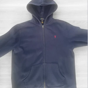 Mörkblå hoodie från Polo Ralph Lauren - Säljer en mörkblå hoodie från Polo Ralph Lauren med dragkedja och huva. Tröjan har en röd broderad logga på bröstet och praktiska fickor framtill. Perfekt för en avslappnad stil. Skulle säga att den är i storlek S förr herr. Hör av dig för information, fler bilder eller annat✅