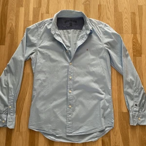 Ljusblå skjorta från Replay Blue Jeans - Säljer en klassisk ljusblå skjorta från Replay Blue Jeans med diskret röd broderad detalj på bröstet. Skjortan har långa ärmar, knappar framtill och vid ärmslut. Perfekt för en stilren look.