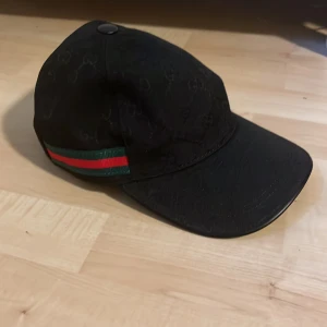 Svart keps - Säljer en svart keps med diskret GG-monogram över hela kepsen. Kepsen har två guldfärgade öljetter på sidan och en klassisk grön-röd rand. Snygg och stilren accessoar. Oanvänd, pris kan diskuteras