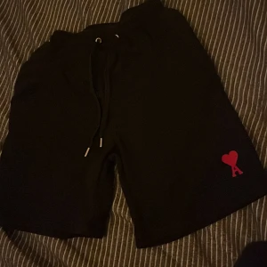 Svarta shorts från Ami Paris med röd broderad logga - Svarta shorts från Ami Paris med snörning i midjan och en röd broderad hjärtlogga med bokstaven A på ena benet. Klassisk och stilren design, perfekta för sommaren.