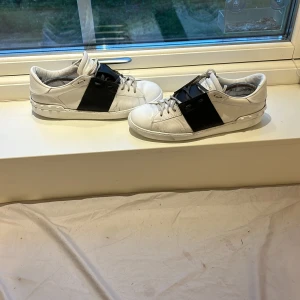 Valentino open svarta - Säljer ett par vita sneakers från Valentino open med en bred svart läderdetalj över sidan. Skorna har rund tå, platt sula och klassisk snörning. Perfekta för dig som gillar stilrena men ändå unika sneakers. Funkar 42 med 