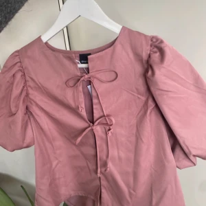 Rosa blus med puffärmar och knytning - Säljer en söt rosa blus med korta puffärmar och två knytband framtill. Blusen har rund halsringning och är tillverkad i ett mjukt bomullsliknande material. Perfekt för vår och sommar!🌸🥂💗🍾Nypris 400kr