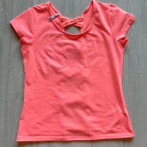 Korallrosa topp med snörningsdetalj i ryggen - Säljer en korallrosa topp med korta ärmar och unik snörningsdetalj i ryggen. Toppen har rund halsringning och är tillverkad i mjuk bomull. Perfekt för dig som vill sticka ut med en cool detalj på ryggen!