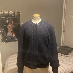 Mörkblå ulljacka med ribbad krage - Säljer en mörkblå jacka i ull med enkel design och ribbad krage. Jackan har lång ärm och rak passform, perfekt för kyliga dagar. Stängs framtill utan synliga detaljer. Passar dig som gillar stilren och klassisk look.