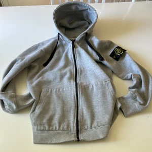 Grå hoodie med dragkedja från Stone Island - Säljer en grå hoodie från Stone Island med dragkedja framtill och svart logotyp-patch på vänster ärm. Tröjan har huva med svarta snören och två fickor framtill. Perfekt för en avslappnad och stilren look. Storlek är XS-S och passar till 165-175