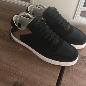 Burberry skor - Snygga svarta sneakers från Burberry med klassiska beige och svarta rutiga detaljer på hälen. Skorna har vit platt sula och svarta snören. Perfekta för dig som vill ha en stilren men ändå unik look.