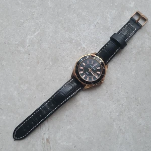 Svart klocka från Dyrberg/Kern - Stilren klocka från det exklusiva danska märket Dyrberg/Kern med svart urtavla och guldfärgad boett. Klockan har svart läderarmband med vita sömmar och klassisk spännesstängning. Urtavlan har guldiga markeringar och detaljer. Gjord av rostfritt stål och kan även visa datumet. Behövs köpas ett nytt batteri.