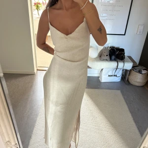 Champagnefärgad långklänning med slits - Säljer en elegant champagnefärgad långklänning med smala axelband och v-ringning. Klänningen har en slits framtill och öppen rygg, vilket ger en stilren och trendig look. Perfekt för sommarens tillfällen!