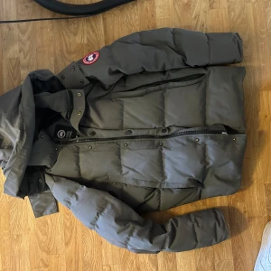 Grå dunjacka från Canada Goose med pälskrage - Säljer en grå dunjacka från Canada Goose med avtagbar pälskrage och klassisk logga på ärmen. Jackan har dragkedja, knappar och flera fickor. Perfekt för kalla dagar och riktigt varm.