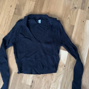 Svart ribbad croppad tröja från H&M Divided - Säljer en svart ribbad croppad tröja från H&M Divided. Tröjan har lång ärm, v-ringning och krage. 