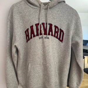 Grå Harvard hoodie - Säljer en klassisk grå hoodie med Harvard-tryck i rött och svart framtill. Tröjan har huva med dragsko och en stor magficka. 