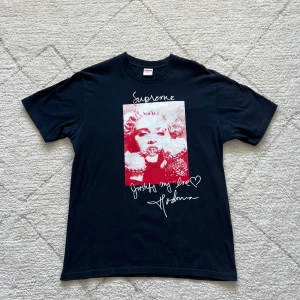 Svart Supreme t-shirt med Madonna-print - Supreme t-shirt i svart med stort rött och vitt print av Madonna på framsidan. Trycket har texten 'Justify my love' och Supreme-loggan ovanför motivet. Klassisk rund hals och korta ärmar. Perfekt statement-plagg för streetwear.