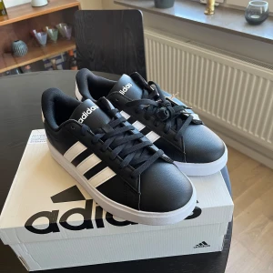 Adidas Grand Court 2.0 sneakers svart/vit - Helt sprillans svarta Adidas Grand Court 2.0 sneakers med vita ränder och vit sula. Klassisk låg modell med snörning och rund tå.  Storlek:  Eu 40   UK 6.5    US 7     Kvitto finns på Mail. Bara att fråga i chatten :)