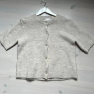 Beige stickad kofta från Gina Tricot - Kortärmad beige stickad kofta från Gina Tricot med rund halsringning och knappar framtill. Perfekt för lager-på-lager och enkel att matcha med olika outfits.