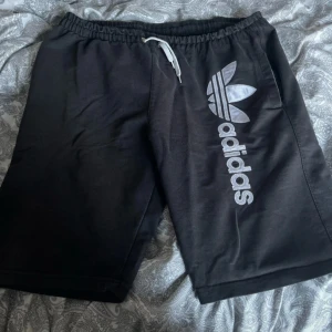 Svarta shorts från Adidas med logga - Svarta shorts från Adidas med stor vit logga och text på vänster ben. Resår i midjan med vit snörning. Perfekta för träning eller chill. Klassisk och sportig stil.