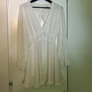 Vit volangklänning med öppen rygg från Dennis Maglic - Superfin vit kortklänning från Dennis Maglic  med volangdetaljer och v-ringning. Klänningen har långärmade ärmar med volang vid ärmslut och en snygg öppen rygg Endast använd en gång.