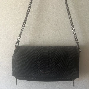 Svart axelväska med kedjerem och ormmönster - Säljer en svart axelväska med snyggt ormmönster och silverfärgad kedjerem. Väskan har dragkedja på baksidan och en stilren design som passar till många outfits. Perfekt storlek för det viktigaste du vill ha med dig. Väskan är använd ca 3 gånger, ingen skada eller repa.