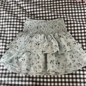 Ljusblå blommig volangkjol från Gina Tricot - Supersöt ljusblå kjol från Gina Tricot med blommigt mönster i rosa, blått och grönt. Kjolen har smock i midjan och dubbla volanger för extra volym. Perfekt för dig som gillar romantisk stil!