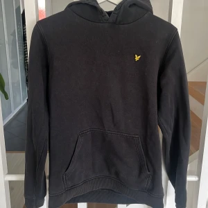 Svart hoodie från Lyle & Scott Junior - Säljer en svart hoodie från Lyle & Scott Junior med klassisk gul logga på bröstet. Tröjan har huva och en stor magficka framtill. Tröjan är välanvänd 