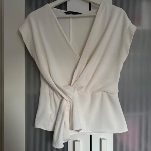Vit omlottopp från Zara - Säljer en stilren vit omlottopp från Zara med knyt i midjan. Toppen har korta ärmar och en djup v-ringning framtill. Perfekt för dig som gillar en elegant och enkel look. Skriv privat för fler bilder 🩷