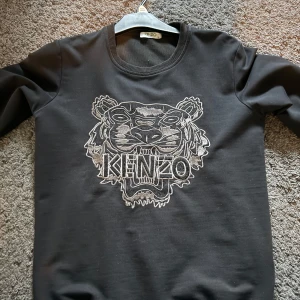 Svart sweatshirt från Kenzo med tigermotiv - Svart sweatshirt från Kenzo med det ikoniska tigermotivet broderat på bröstet. Tröjan har rund halsringning och långa ärmar. Perfekt för dig som vill ha en stilren men ändå statement tröja.
