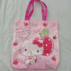Hello Kitty rosa shoppingväska - Supergullig rosa shoppingväska med Hello Kitty-tryck och stora jordgubbar och äpplen. Väskan har två starka rosa handtag och är perfekt för att bära böcker eller småsaker. Söt design med hjärtan och små detaljer.