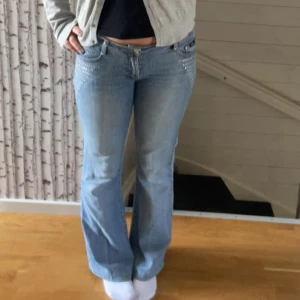 Ljusblå bootcut jeans med nitar - Säljer ett par ljusblå jeans med bootcut passform och snygga nitar vid fickorna. Jeansen har normal passning och klassisk femficksdesign. Perfekta för dig som gillar en retroinspirerad stil. Har ett litet hål mellan bena men går lätt att fixa