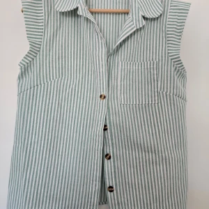 Randig skjorta med knappar och krage - Snygg skjorta med gröna och vita vertikala ränder, klassisk krage och knäppning framtill. Skjortan har en bröstficka och är ärmlös.