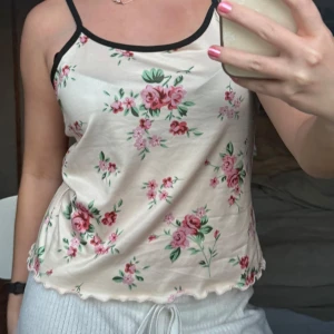 Blommig linnetopp med svarta kanter - Söt linnetopp med blommigt mönster i rosa och grönt på ljus botten. Toppen har tunna axelband och svarta kanter runt halsringningen. Perfekt för varma dagar och enkel att matcha med jeans eller kjol.