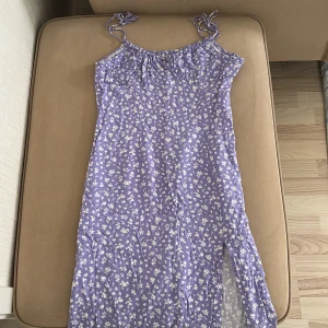 Ljuslila blommig långklänning med slits - Säljer en söt ljuslila långklänning med vita småblommor och smala axelband som knyts upptill. Klänningen har en slits på sidan och är perfekt för varma dagar. Materialet känns lätt och luftigt. Den går över knäna på mig som är 170.