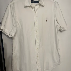 Vit kortärmad skjorta från Ralph Lauren - Klassisk vit skjorta från Ralph Lauren med korta ärmar och knappar framtill. Skjortan har den ikoniska broderade loggan på bröstet och är tillverkad i bomull. Perfekt för en stilren och enkel look. Använd 3 gånger, inga defekter den lilla fläcken vid märket är en ångfläck som ej syns längre.