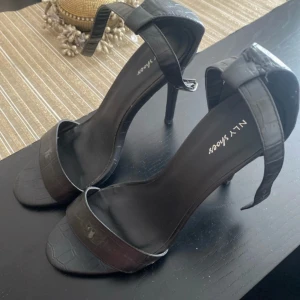 Svarta klackskor med krokodilpräglat mönster från NLY Shoes - Säljer ett par svarta klackskor från NLY Shoes med trendigt krokodilpräglat mönster. Skorna har öppen tå, smal rem över tårna och bredare rem runt vristen med spänne. Perfekta för att lyfta din outfit.