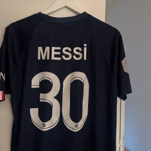 PSG fotbollströja Messi 30 - Säljer en mörkblå PSG fotbollströja med Messi och nummer 30 på ryggen. Tröjan har vita och röda detaljer, PSG-logga på bröstet och sponsortryck framtill. Kortärmad modell, perfekt för fotbollsfans.