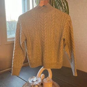 Grå stickad tröja med kabelmönster - Säljer en grå stickad tröja med klassiskt kabelstickat mönster och ribbade muddar. Tröjan har lång ärm och hög hals, perfekt för kyligare dagar. Snygg och enkel att matcha till olika outfits.