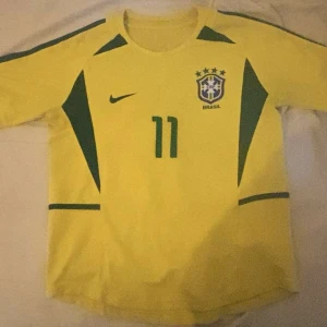 Brasilien fotbollströja Ronaldinho #11 - Gul Brasilien fotbollströja från Nike med gröna detaljer och nummer 11 på bröstet och ryggen. Ronaldinho står tryckt på ryggen. Klassisk landslagströja med korta ärmar och rund hals. Storleken är M men passar mig som är biffig L-XL