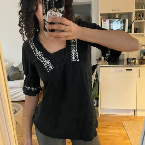 Svart blus med broderi från KappAhl - Säljer en superfin svart blus från KappAhl med vackra vita broderier längs halsringningen och ärmarna. Blusen har en v-ringad design och trekvartsärmar som ger en avslappnad men stilren look. Perfekt för både vardag och fest!