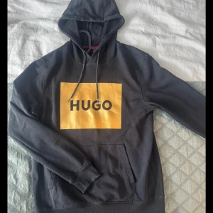 Hugo hoodie - Säljer en svart hoodie från Hugo med stor gul logga på bröstet. Tröjan har huva med dragsko och en klassisk känguruficka framtill. Perfekt för en avslappnad och stilren look.