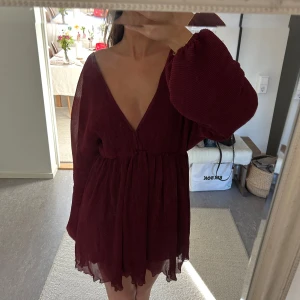 Vinröd kortklänning med v-ringning och ballongärmar - Vinröd kortklänning med djup v-ringning, långa ballongärmar och markerad midja. Klänningen har öppen rygg med knytband och är tillverkad i ett lätt, transparent tyg med fint fall. Aldrig använd med prislapp kvar!