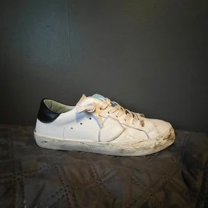 Vita sneakers från Philippe Model - Säljer ett par vita sneakers från Philippe Model med svarta detaljer på hälen och klassisk snörning. Skorna har en platt sula och rund tå, samt diskreta sömmar och logga på baksidan. Perfekta för en stilren och avslappnad stil. Priset går alltid att diskutera!