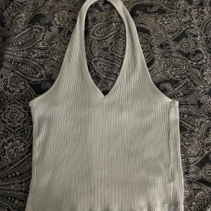 Ljusgrå ribbad halternecktop från NA-KD - Säljer en ljusgrå ribbad halternecktop från NA-KD. Toppen har en djup v-ringning och är ärmlös, perfekt för varma dagar. Mjuk och stretchig i materialet för en bekväm passform.