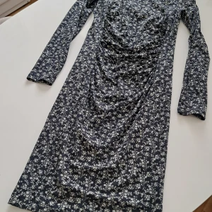 Blommig långärmad klänning från Twin-Set Simona Barbieri - Supersnygg svart klänning med vita blommor från Twin-Set Simona Barbieri. Långärmad modell med rynkade detaljer framtill och figurnära passform. Tillverkad i mjuk viskosblandning med stretch för extra komfort.