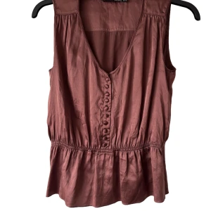 Auburn sidenliknande blus med knappar - Säljer en rostbrun ärmlös blus med v-ringning och dekorativa knappar framtill. Blusen har en markerad midja med lätt volang nedtill och är tillverkad i ett mjukt, lätt material. Varumärke: BGN. Perfekt för varmare dagar!