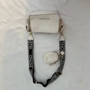 Vit axelväska från Valentino med logoband - Snygg vit axelväska från Valentino med bredt svart axelband med vit logga. Väskan har guldfärgade detaljer och en mindre matchande ficka som sitter fast på bandet. Perfekt för att bära det viktigaste med stil.