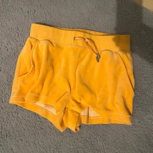 Orange velourshorts från Juicy Couture - Säljer ett par mjuka och sköna orange velourshorts från Juicy Couture med dragsko i midjan och broderad JC-logga på ena benet. Perfekta för sommarens varma dagar eller som mysplagg hemma.