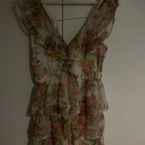 Blommig volangklänning från Nelly Eve - Klänning från Nelly Eve med volangdetaljer och v-ringning. Klänningen har flera lager i ett skirt material och dragkedja i sidan. Perfekt för sommaren.