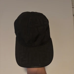 Svart keps med mönster från Fendi - Säljer en svart keps från Fendi med diskret mönster i tyget. Klassisk form med böjd skärm och justerbar passform. Perfekt accessoar för en stilren look.