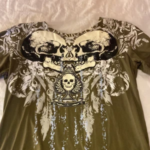 Olivgrön t-shirt med döskalletryck - Cool olivgrön t-shirt med stort tryck av två döskallar och ett spaderess med döskallemotiv framtill. T-shirten har korta ärmar och rund halsringning. Perfekt för dig som gillar unika och edgy plagg.