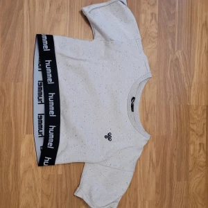 Vit croppad topp från Hummel - Säljer en vit croppad topp från Hummel med svart resår i nederkant där Hummel-loggan upprepas. Toppen har korta ärmar och en liten Hummel-logga på bröstet. Perfekt för en sportig look!